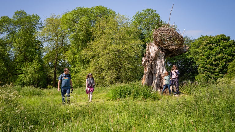 A family plays near the sun sculpture in the centre of the parkland, Lacock Abbey. It is summer, the sun is shining and the children smile towards the camera.
